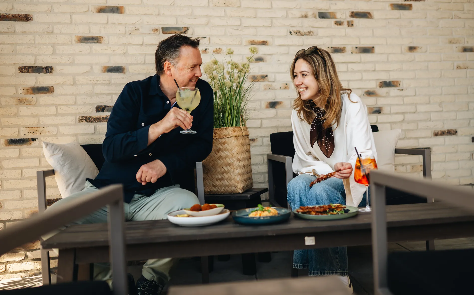 Een man en een vrouw die gezellig praten en lachen tijdens een borrel met hapjes en cocktails op het loungeterras tegen een lichte bakstenen muur.